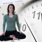 Die Meditationslehrerin Stefanie Seher sitzt mit geschlossenen Augen im Meditationssitz. Den Hintergrund bilden ein großes Kalenderblatt und das Ziffernblatt einer Uhr.
