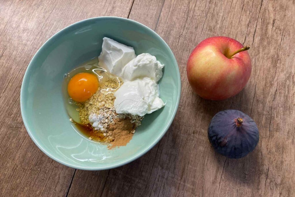 Zutaten für gebackenen Porridge: Haferflocken, Joghurt, Ei, Zimt, Backpulver und Ahornsirup in einer Schale, daneben ein Apfel und eine Feige